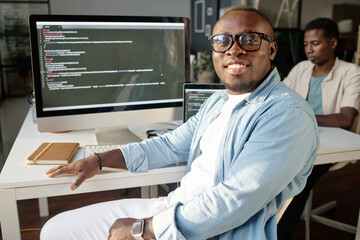 Medium closeup portrait of joyful programmer sitting at table and looking at camera