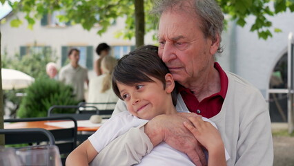 Fototapeta premium Grandfather holding his grandson at an outdoor cafe, both smiling and enjoying each other's company, surrounded by greenery, capturing a warm and loving family moment