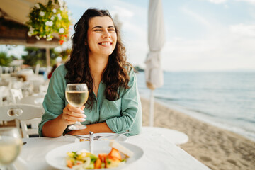 Young caucasian woman eating greek salad and drinking wine by the sea
