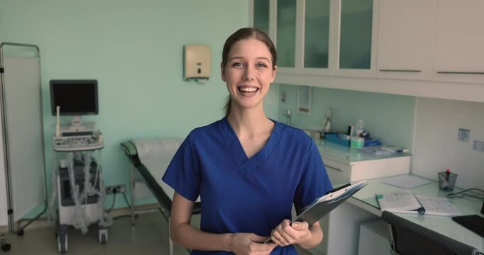 Happy young woman intern, licensed practical nurse or assistant in blue uniform poses in hospital. Clinical support, healthcare facility administrative staff member, medical laboratory worker portrait