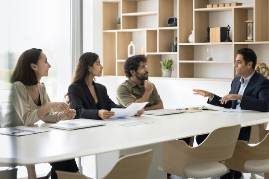 Group of multiracial students trainees of different age gender attend lecture seminar listen to Indian male coach mentor at meeting room. Project manager speak to teammates present benchmarking report