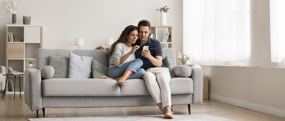 Young millennial couple of homeowners sitting on cozy couch in modern home interior, using online application, service, domestic Internet technology on mobile phone together. Wide full length shot