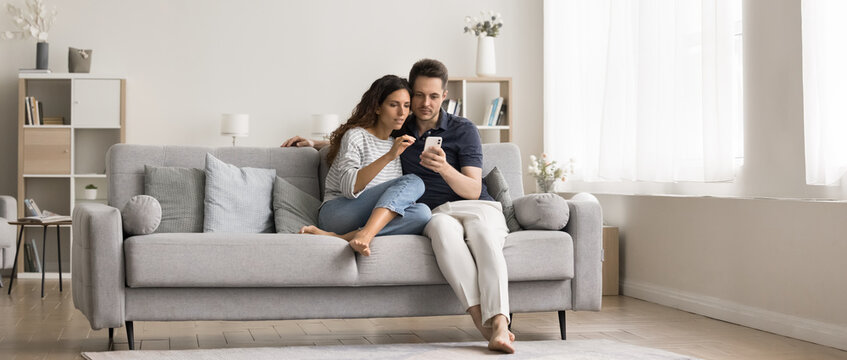 Young millennial couple of homeowners sitting on cozy couch in modern home interior, using online application, service, domestic Internet technology on mobile phone together. Wide full length shot