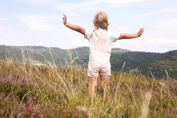 Fototapeta premium Cute little girl standing on meadow near mountains, back view