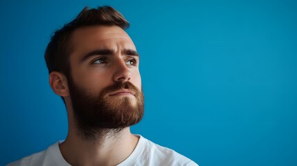 Fototapeta premium Thoughtful Young Man with Beard Looking Up Against Blue Background