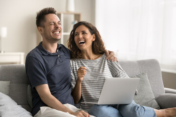 Happy joyful husband and wife sitting on couch with gadget, holding laptop, using Internet communication, enjoying wireless technology, online service, leisure at home, laughing, having fun