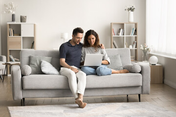 Millennial couple using Internet service, application on laptop, sitting on couch together, watching online content, enjoying leisure, wireless technology in comfortable home interior