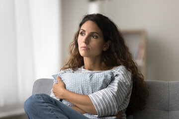 Serious pensive pretty Latin woman sitting on sofa at home, looking away in deep thoughts,...