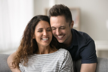 Fototapeta premium Joyful positive married couple having fun at home, enjoying leisure. Cheerful handsome husband hugging wife from behind sitting at comfortable couch with closed eyes, laughing
