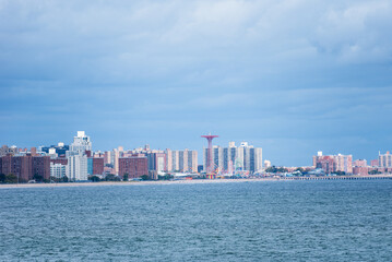 Fototapeta premium The skyline of Brooklyn emerges from the horizon, seen from a ship sailing through Gravesend Bay. The buildings stand in contrast against the cloudy sky and the steel-colored waters of the bay.