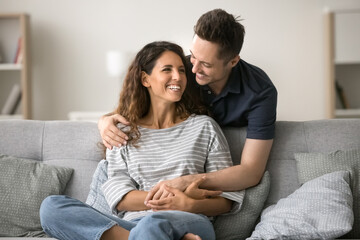 Cheerful attractive couple in love enjoying leisure at home, hugging at comfortable couch, talking, laughing. Happy handsome husband embracing wife from behind