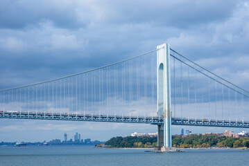 The Verrazzano-Narrows Bridge stretches across the Upper Bay near New York City, its steel arches silhouetted against a cloudy sky. The towering structure stands as a monumental gateway to the city.