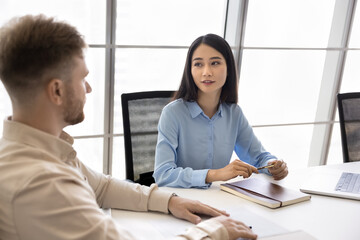 Obraz premium Two diverse business colleagues talking at office meeting table. Young Asian employee, manager woman discussing work project, partnership, teamwork with male boss, partner