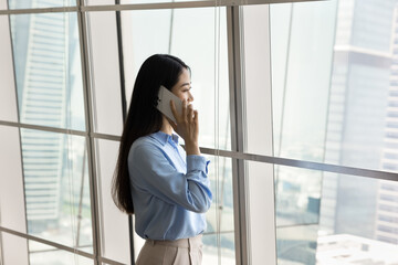 Thoughtful young Asian business woman talking on cellphone in office space, standing at large window, looking away at city view, thinking on conversation, commercial communication