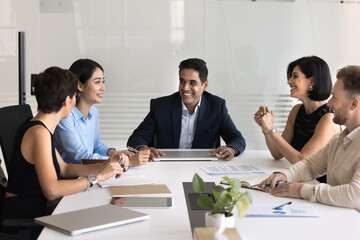 Happy middle aged Indian company director man holding meeting with diverse employees. Multiethnic managers talking at large office table, brainstorming on teamwork creative strategy, laughing