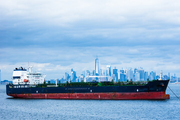 Obraz premium A cargo ship rests anchored in the bay, dominating the foreground. In the distance, the steel-blue skyline of New York City looms under a cloudy sky, blending into the moody, overcast atmosphere.