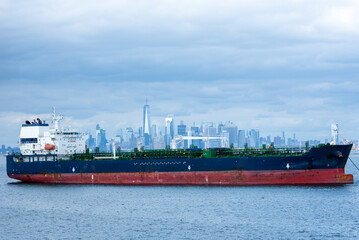 Fototapeta premium A cargo ship rests anchored in the bay, dominating the foreground. In the distance, the steel-blue skyline of New York City looms under a cloudy sky, blending into the moody, overcast atmosphere.