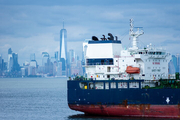 Obraz premium The ship's stern takes focus as it glides through the bay, with New York City's skyline blurred in the background. The cloudy sky casts a soft, steel-blue tone over the entire scene.
