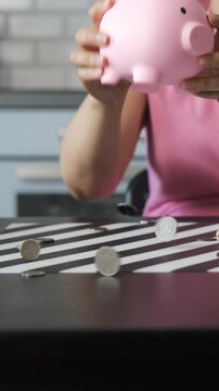 Vertical video. Young woman clears a piggy bank, spreading coins across a paper-covered table