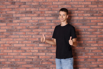 Teenage boy wearing black t-shirt and showing thumbs up near brick wall