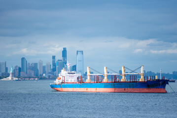 A cargo ship rests anchored in the bay, dominating the foreground. In the distance, the steel-blue skyline of New York City looms under a cloudy sky, blending into the moody, overcast atmosphere. © Mariusz