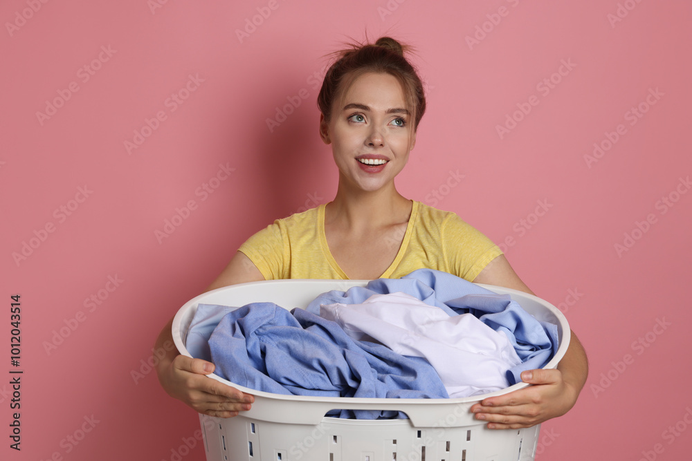 Poster Happy young housewife with basket full of laundry on pale pink background