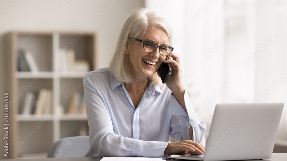 Wall mural happy businesswoman typing on laptop keyboard and talking on smartphone, working seated at workplace