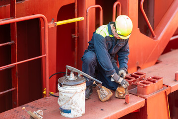 A crew member is crouched on the deck of a merchant ship, applying grease to container securing...