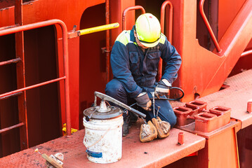 A crew member is crouched on the deck of a merchant ship, applying grease to container securing equipment. Focused on ensuring the safe and smooth operation of the gear during the voyage.