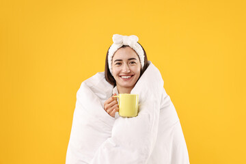 Happy young woman with cup wrapped in blanket on yellow background