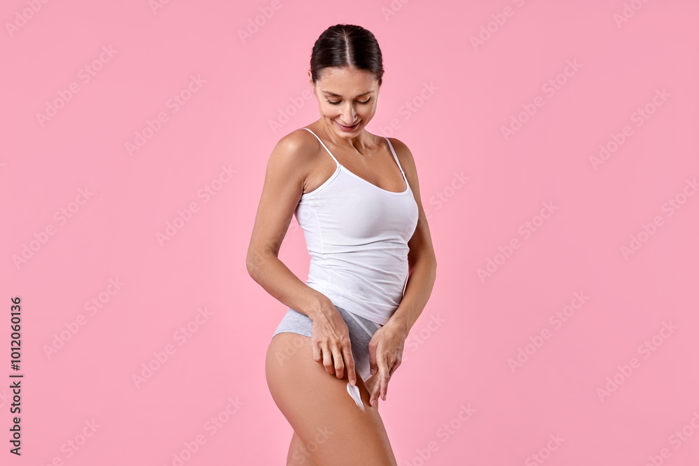 Wall mural Beautiful woman applying cream onto leg against pink background. Body care