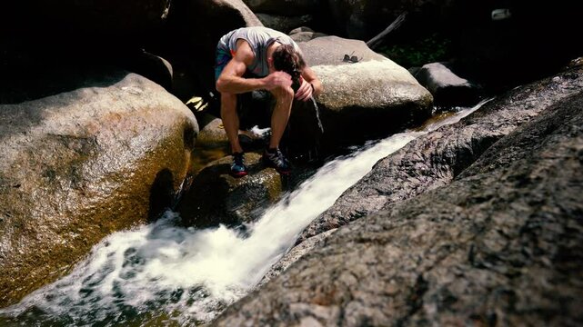 A man sitting on a large stone rinses his head with cold river water