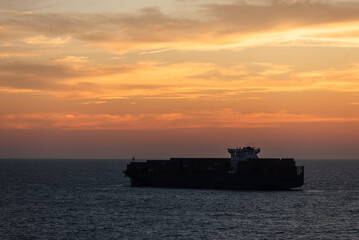 The calm silhouette of a massive container ship gliding across the smooth sea, enveloped by the golden hues of the sky as the sun bids farewell to the day.