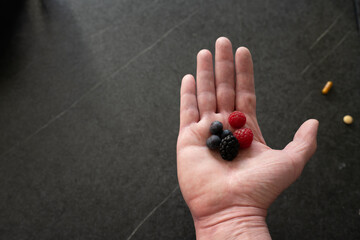 Hand holding some berries and presenting them. Some pills, like a capsule and a pill, can be seen lying on the table. Pills blurry. Black background, white space.