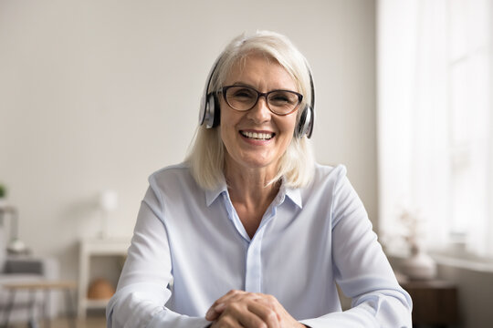 Smiling middle-aged woman sitting at table wear headphones engaged in video call or online meeting, take part in virtual remote communication, provide consultation, lead on-line class or webinar event