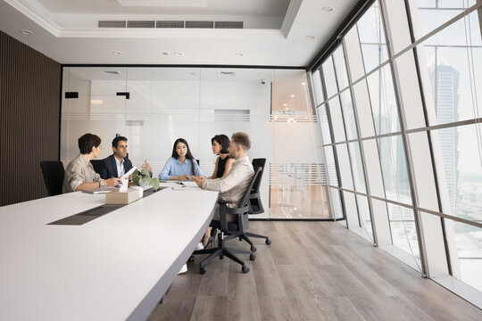 Diverse group of company stakeholders talking at large table in contemporary office workspace. Business colleagues discussing teamwork in stylish meeting room interior. Wide shot with copy space