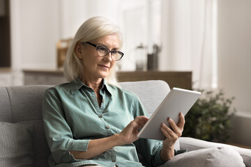 Pretty mature female wear eyeglasses holding digital tablet device chatting with family, web...