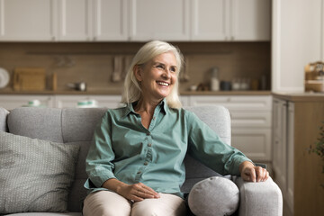 Smiling older female resting on comfortable sofa in studio apartment, looks into distance, deep in...