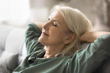 Close up serene European elderly woman reclining back with her eyes closed, enjoy moment of peace and tranquillity, relaxing comfortable in cozy environment leaned on couch at home. Calmness, repose