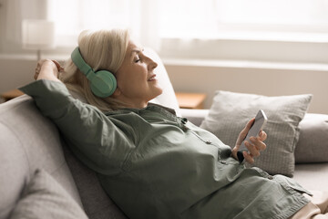 Older woman resting leaned on couch with serene expression, wearing wireless headphones, holding...