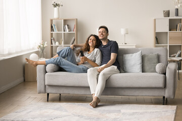 Obraz premium Cheerful millennial husband and wife in love relaxing on sofa together, hugging, watching TV show, sitcom series, movie, sport game broadcasting, laughing, enjoying leisure in modern home interior