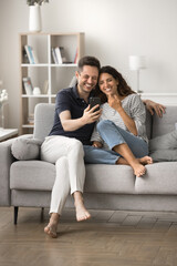 Positive romantic couple in love using smartphone for domestic Internet communication, taking selfies photo at in cozy home interior, resting on sofa, enjoying leisure. Vertical shot