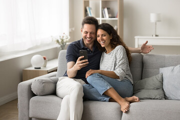 Cheerful married couple using application on mobile phone for Internet communication, sitting together on couch at home, having fun, smiling, laughing, watching online content on smartphone