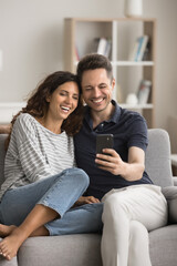 Obraz premium Happy young couple in love using mobile phone for video call, online communication, sitting close together on sofa, holding cellphone, looking at screen, smiling, taking selfie. Vertical shot