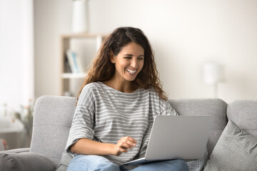 Happy beautiful Latin woman sitting on comfortable sofa with computer, typing on laptop, chatting, online, smiling, laughing, working on freelance business project