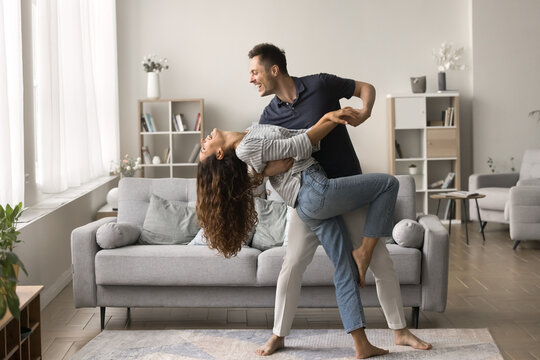 Happy active married couple dancing in stylish living room interior, enjoying activity at home together. Positive strong husband embracing wife in romantic social dance with passion