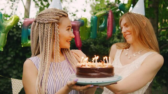 Two cheerful and vibrant girlfriends with red hair and dreadlocks are joyfully celebrating a birthday outdoors, enjoying delicious cake with candles and creating delightful memories together