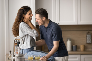 Happy joyful couple having fun in home kitchen, sitting on counter, drinking morning beverage, talking, smiling, laughing, enjoying domestic leisure, romantic weekend together