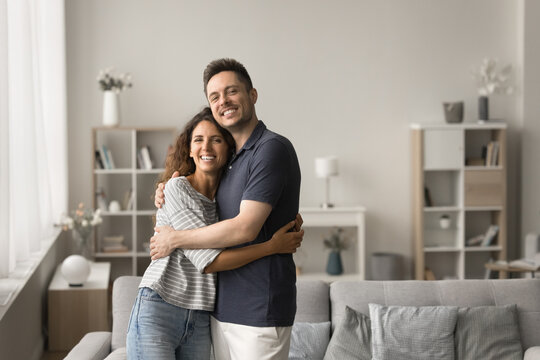 Cheerful attractive young married couple of new homeowners portrait. Happy husband and wife standing in modern home interior, hugging, smiling, laughing, looking at camera