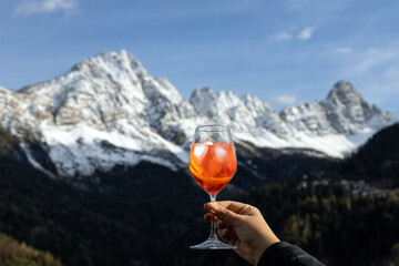 Hand holding a glass of Spritz cocktail, enjoying panoramic view of snowy Dolomites mountains, Italy